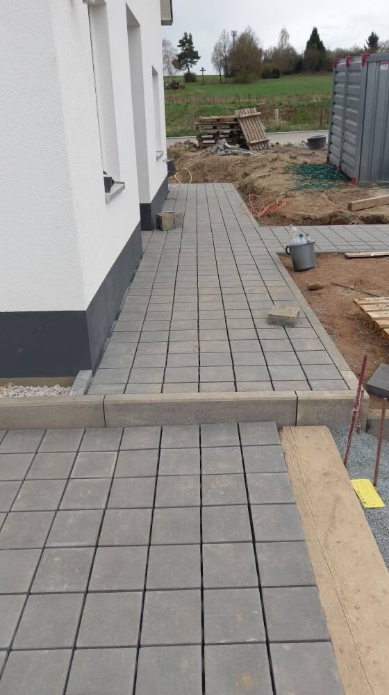 Außenansicht: Gehweg aus grauen Pflastersteinen vor weißem Haus; Baukisten im Hintergrund.