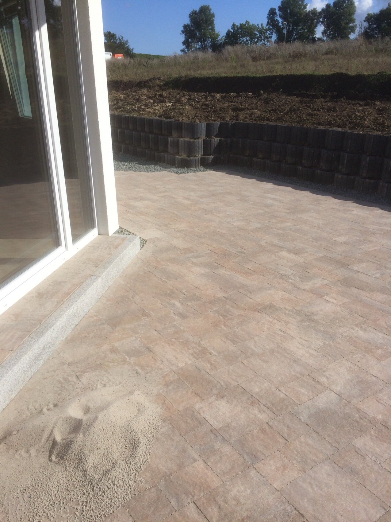 Terrasse mit beigen Pflastersteinen vor Glas-Schiebetür, Baustellenhügel und Stützmauer