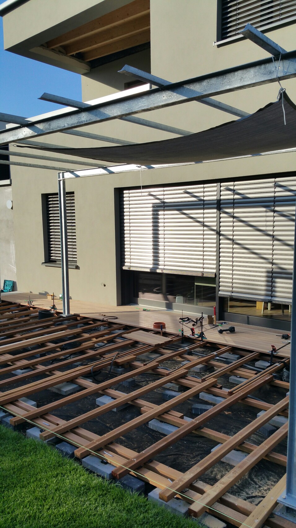 Holzterrassen-Unterbau vor modernem Haus; Metallrahmen mit Sonnensegel, Werkzeuge liegen bereit.