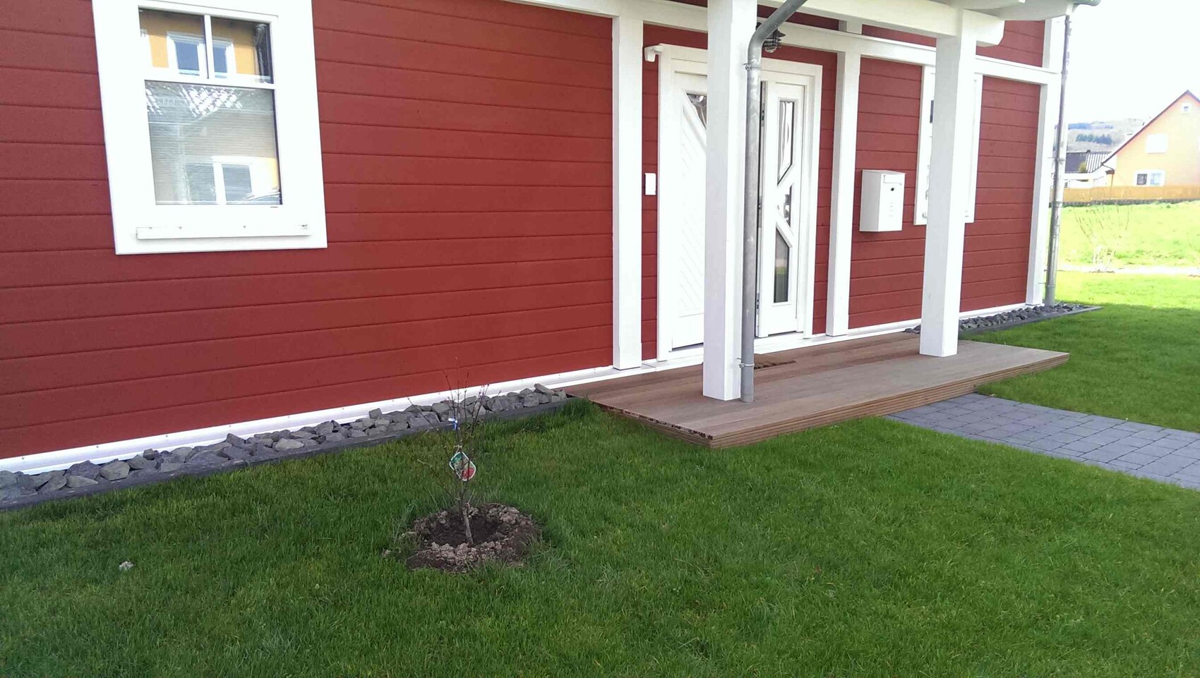 Rotes Holzhaus mit weißer Veranda, Tür und Holzdeck-Eingang, grünem Rasen und kleinem Baum