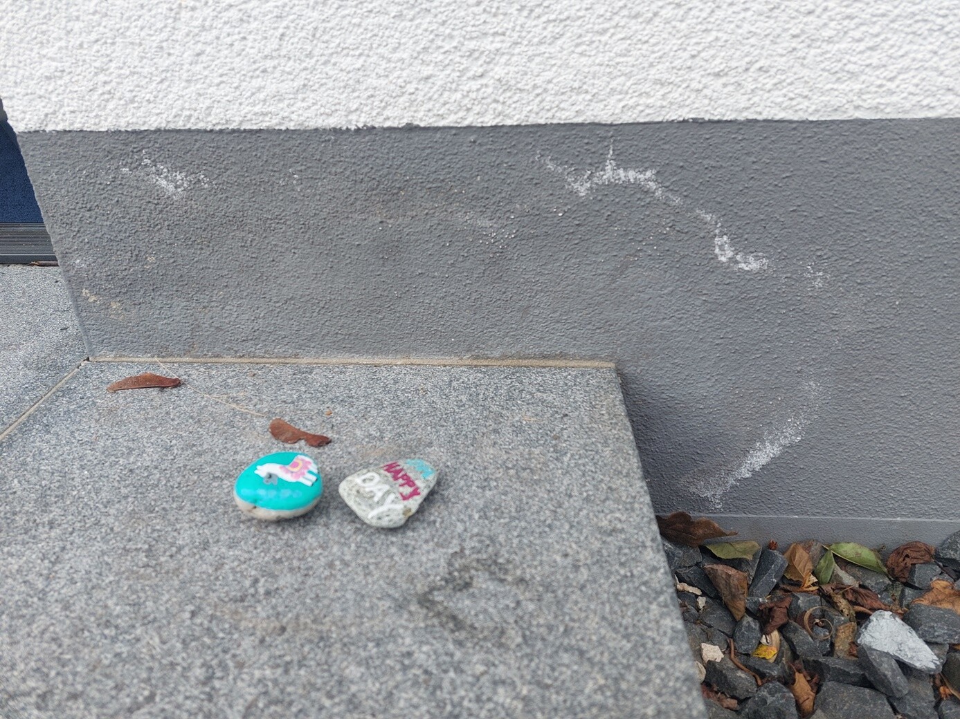 Zwei bemalte Kieselsteine auf einer Betonstufe vor einer grauen Wand; Herbstblätter drumherum.