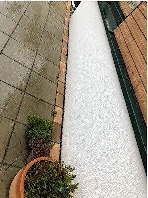 Blick auf Terrasse mit grauen Fliesen, weißer Wand und Holzverkleidung; Topfpflanze im Vordergrund.