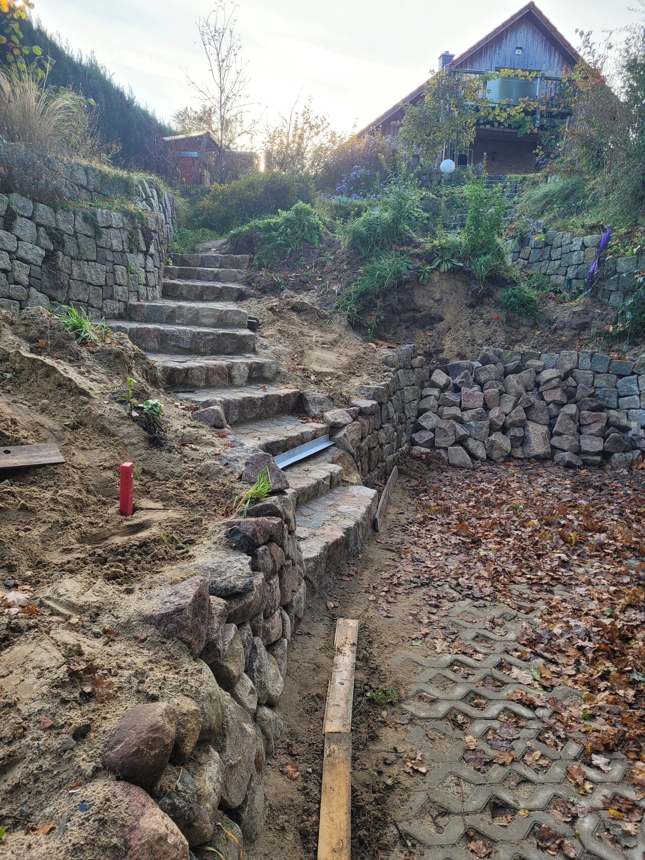 Naturstein-Treppen und Stützmauern in einem Garten; Erde, Laub, Pflanzen, Haus im Hintergrund.