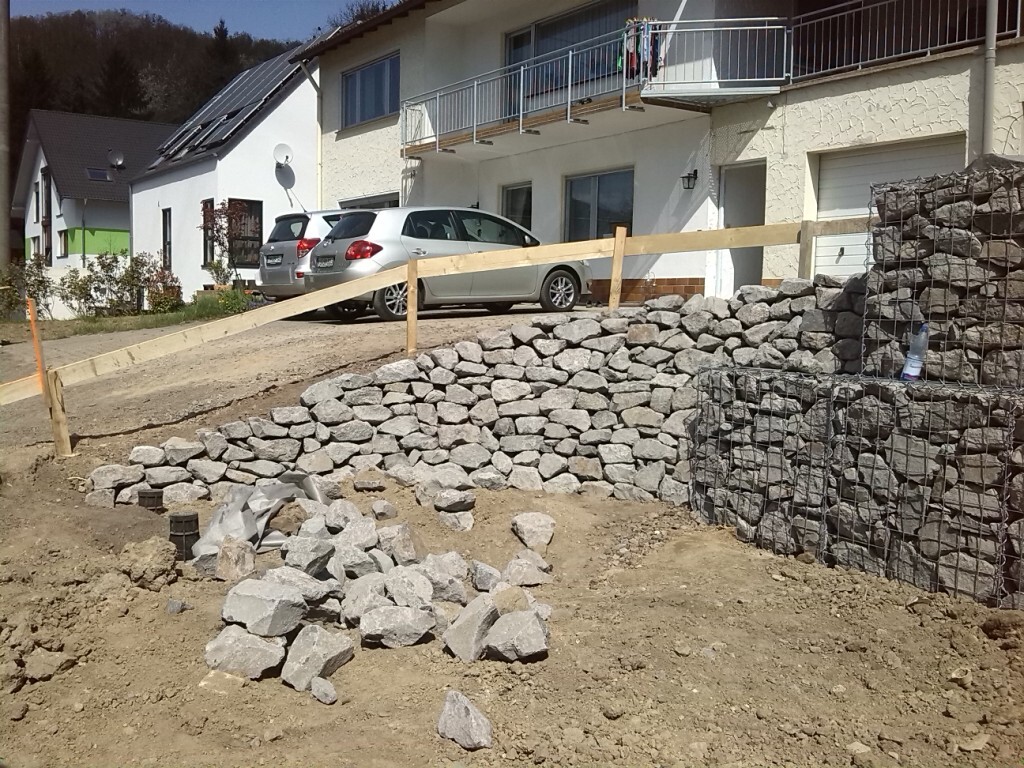 Baustelle vor Wohnhaus: Gabionen-Stützwand, Holzgeländer, Autos auf Einfahrt, Steinehaufen.