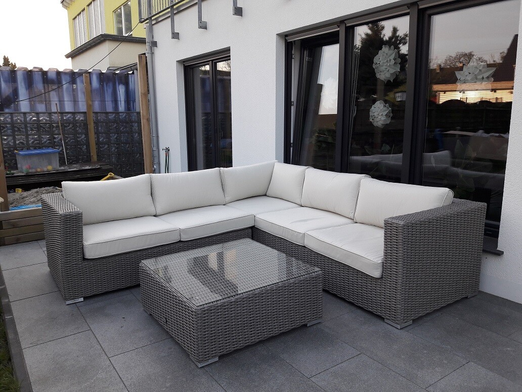 Gartenlounge aus grauem Rattan mit hellen Sitzkissen und Glas-Couchtisch auf Terrasse.