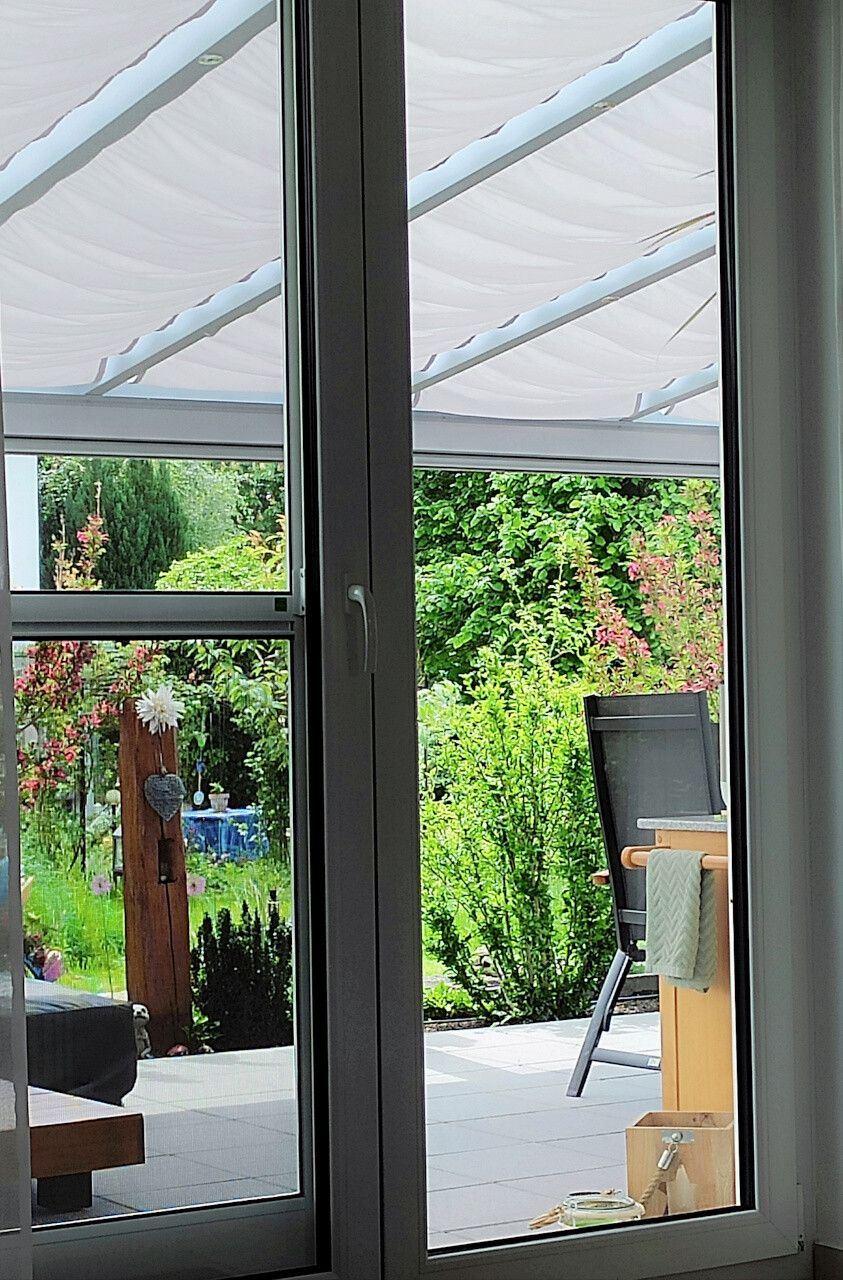 Blick durch eine Glasschiebetür auf einen gepflegten Garten mit Terrasse und Sitzmöbeln.