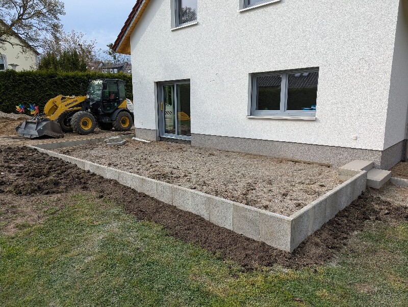 Außenansicht: erhöhte Kies-Terrasse aus Betonblöcken am Haus, Bagger links.[/alt]