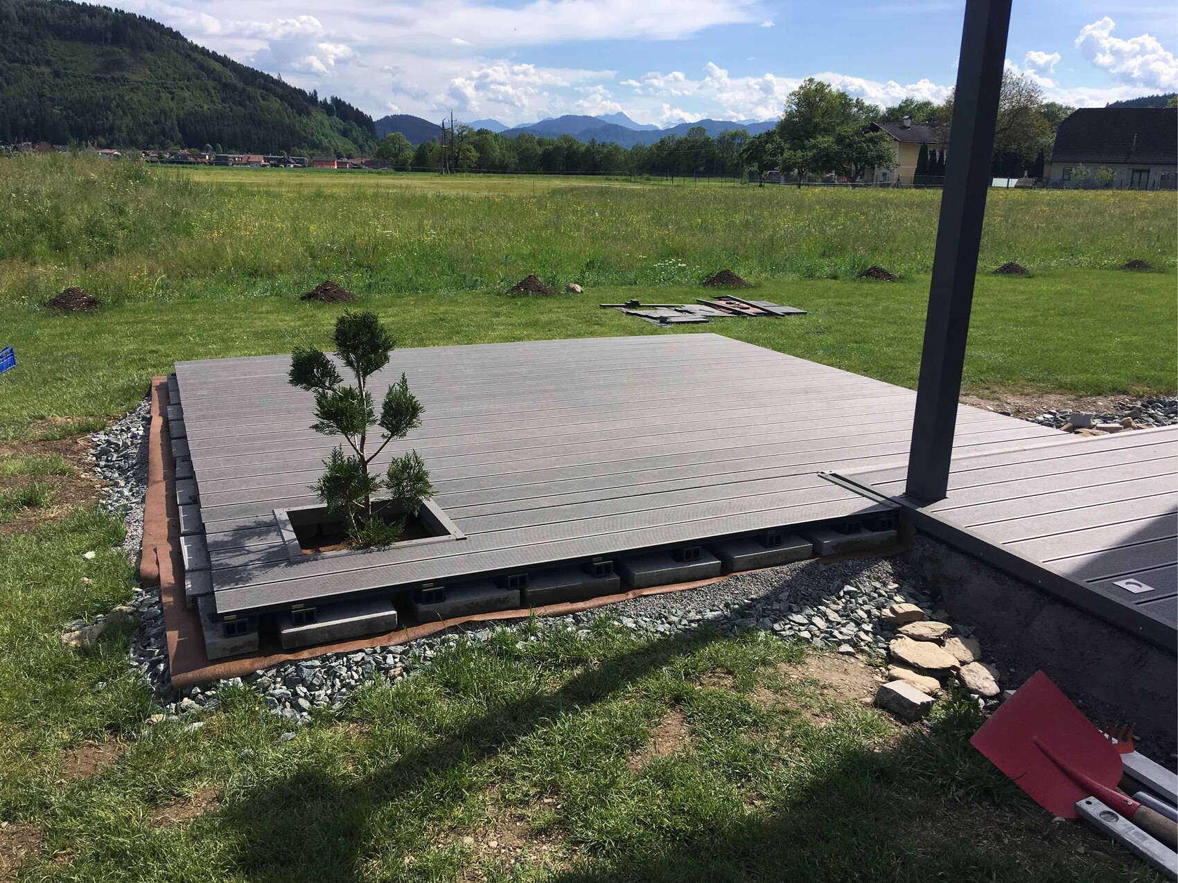 Holzterrasse auf Stelzblöcken im Bau; Baum in Deckenaussparung; grüne Wiesen und Berge.