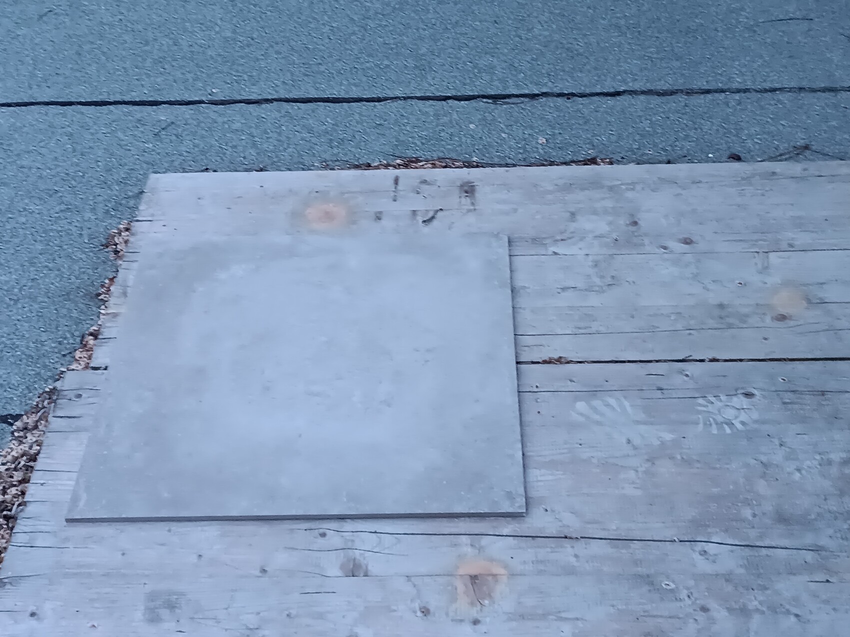 Grau quadratische Metallplatte liegt auf abgenutzten Holzplanken neben einem Asphaltweg.