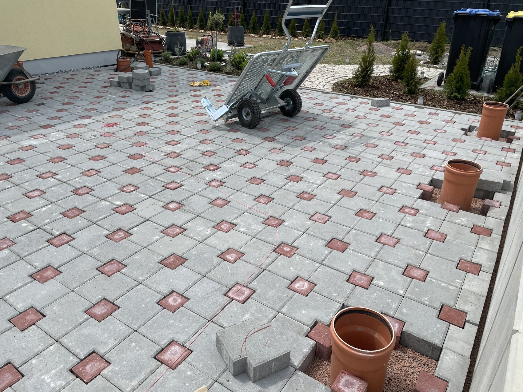 Gepflasterte Terrasse aus grauen Platten mit roten Quadraten; Bauwerkzeuge, Wagen und Tonrohre.