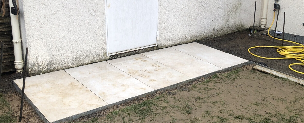Beige Fliesen-Terrasse vor weißer Tür, daneben Erde; gelber Schlauch liegt rechts.
