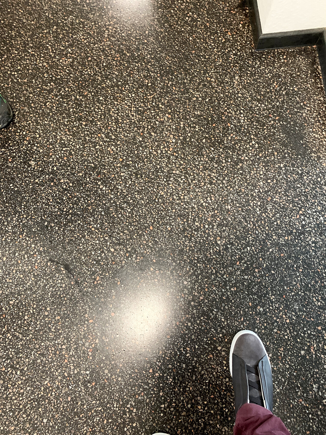 Dunkler Terrazzo-Boden mit bunten Flecken; sichtbarer grauer Sneaker neben burgunder Hose.