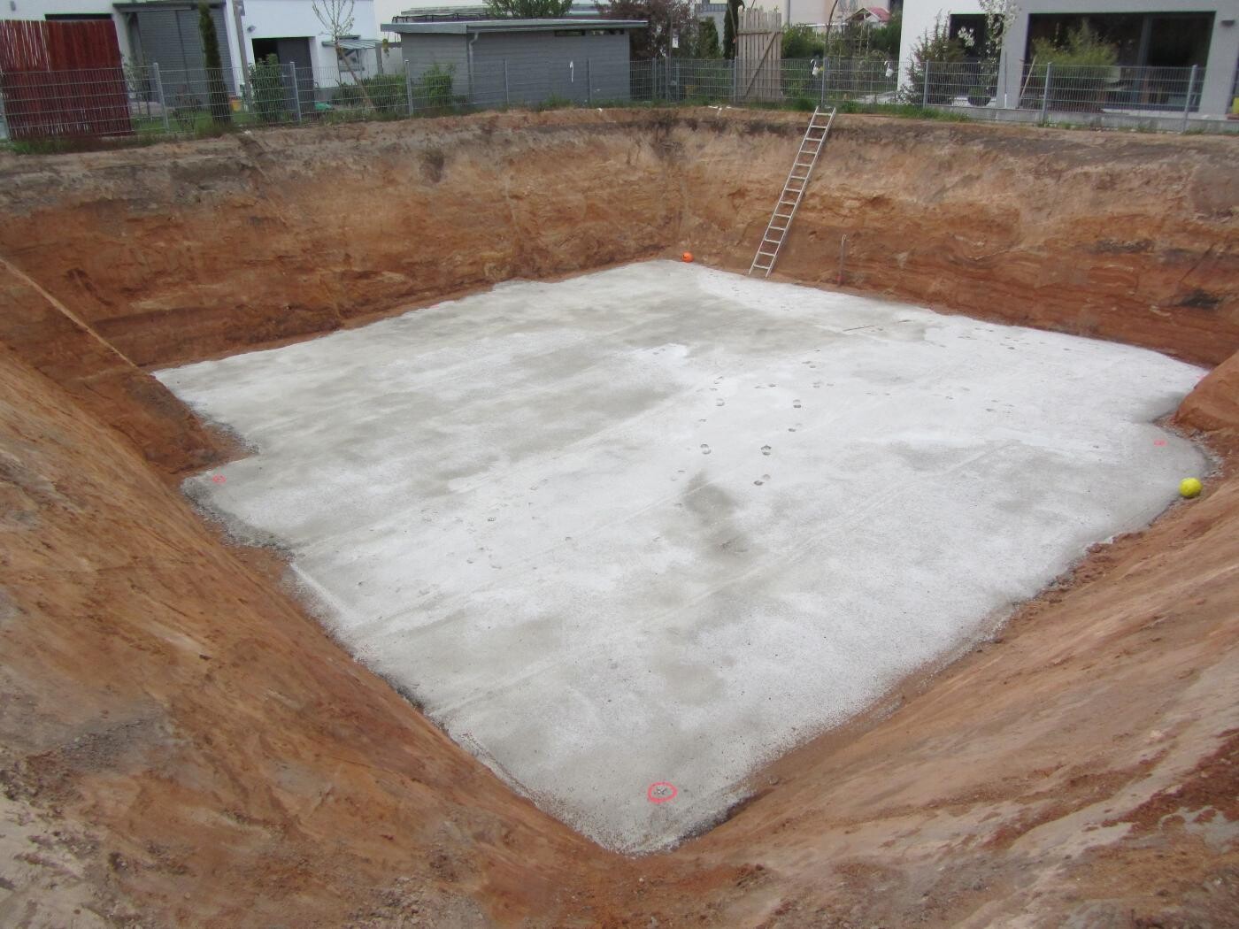 Große Baugrube mit Betonboden, Leiter an der Wand, Umzäunung und Häuser im Hintergrund.