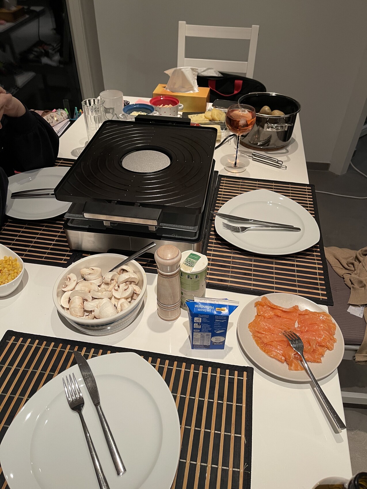 Tisch mit elektrischem Raclette-Grill, Lachs, Pilze, Mais, Wein und Besteck.