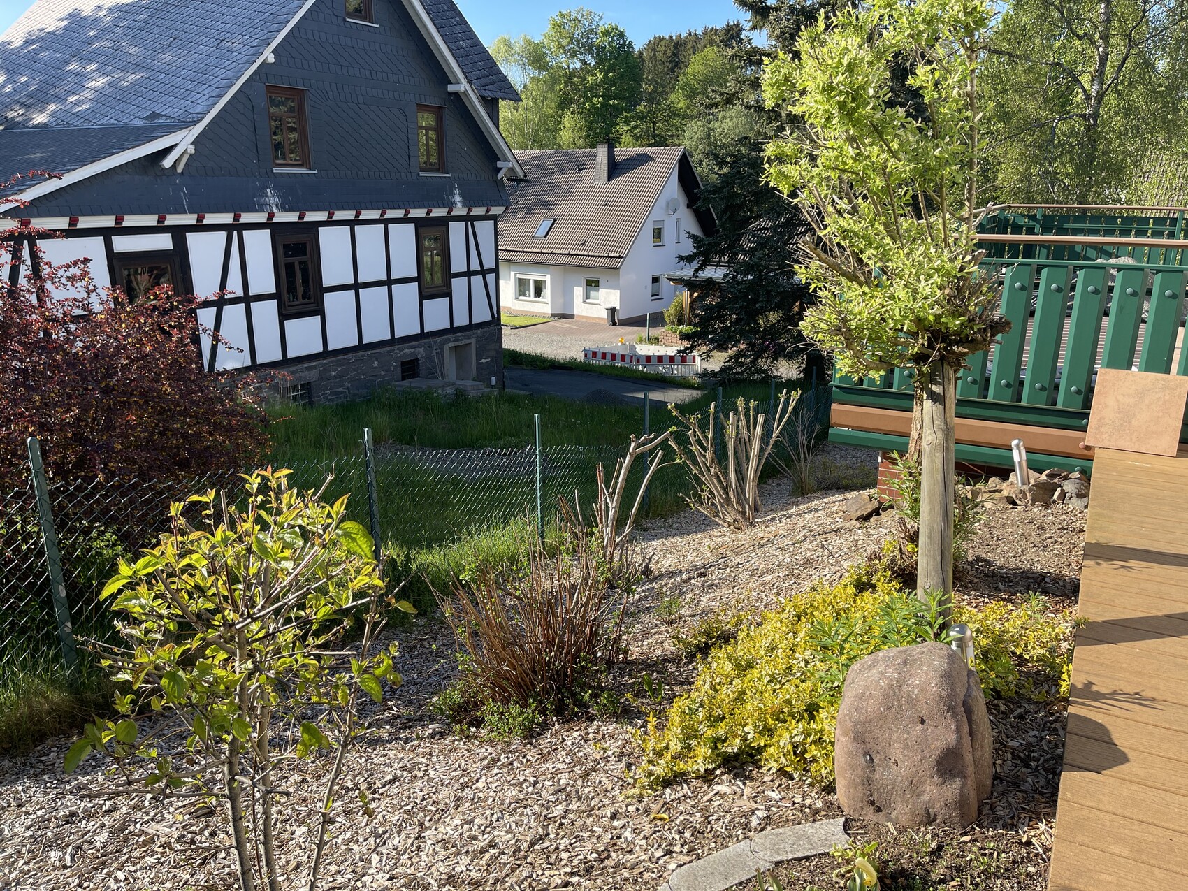 Gartenansicht mit Fachwerkhaus links, Kiesfläche, Baumpflanzen und Holzdeck rechts.