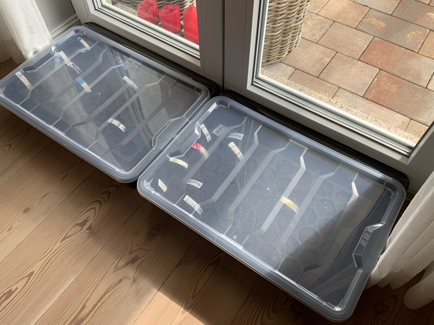 Zwei transparente Kunststoff-Aufbewahrungsboxen mit Fächern stehen auf Holzboden nahe einer Glastür.