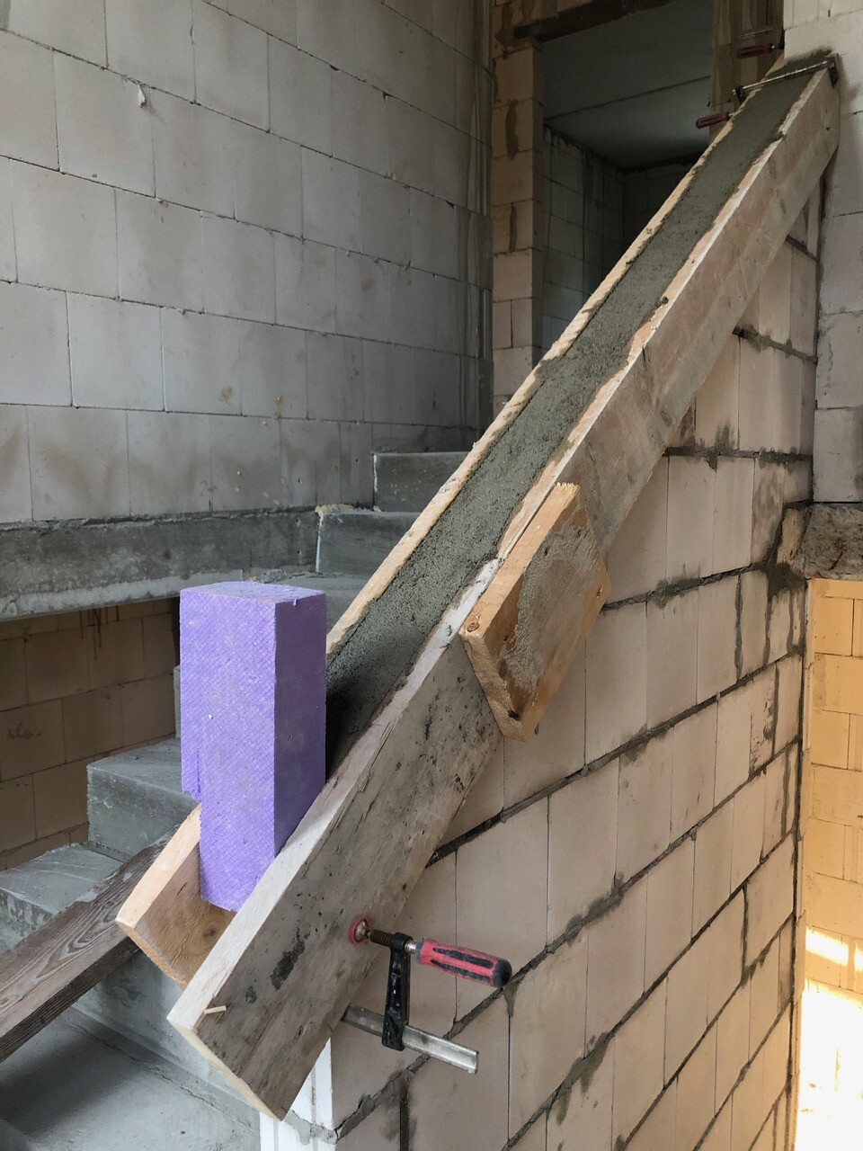 Unfertige Baustellen-Treppe aus Holz an einer Betonwand, Klemmen, violetter Schaumstoffblock.