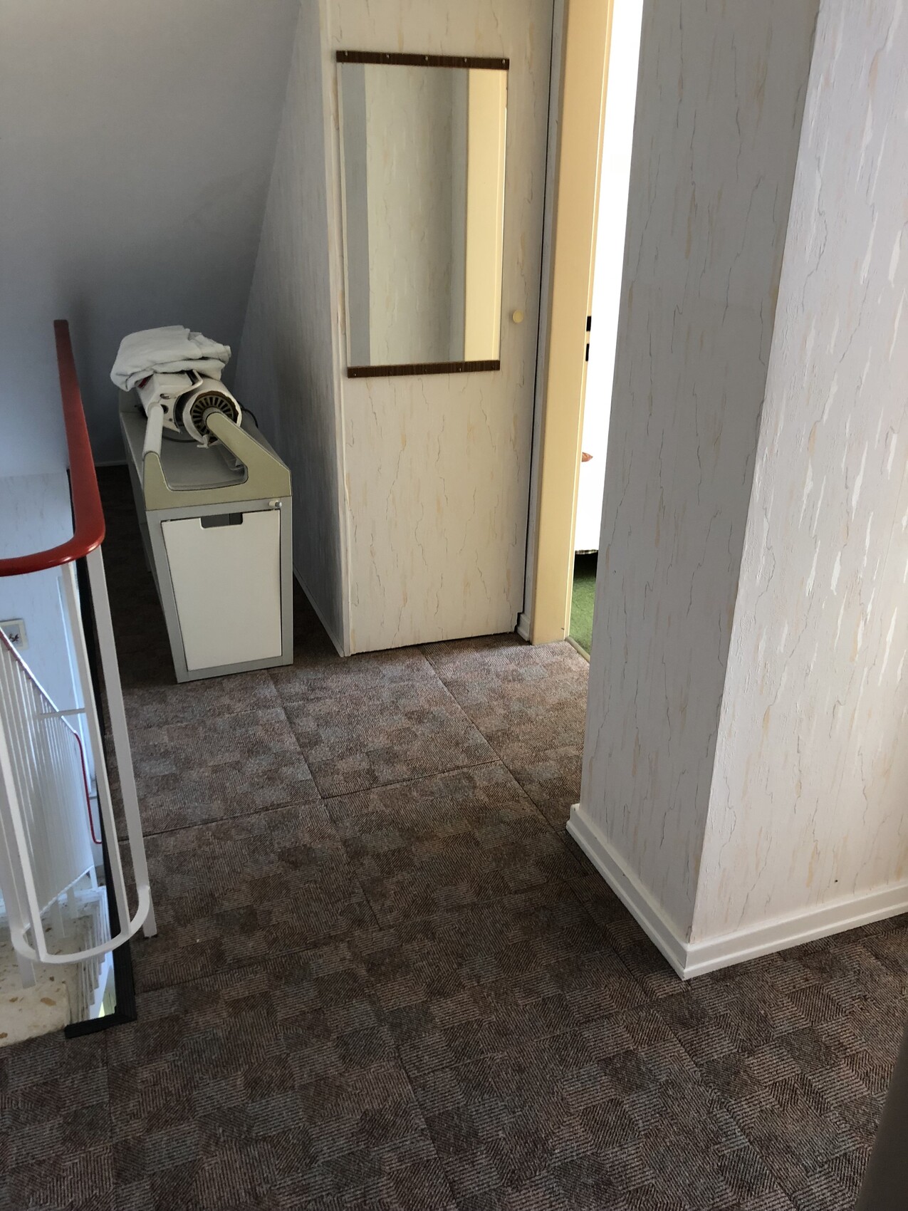 Korridor mit braun gemustertem Teppich, Spiegel an der Wand, ein kleines Gerät auf einem Schrank, Tür nach draußen rechts.