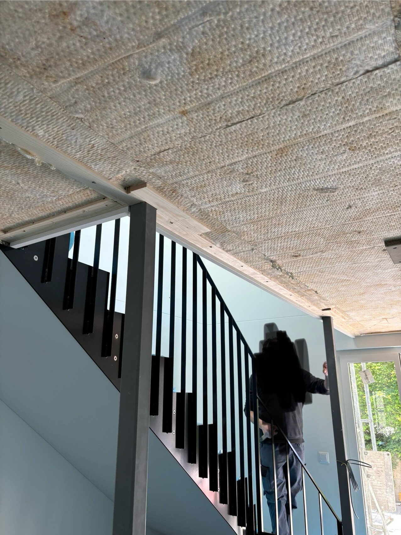 Innenraum mit Treppe und schwarzem Geländer, sichtbare Isolierdecke, Person am Geländer.