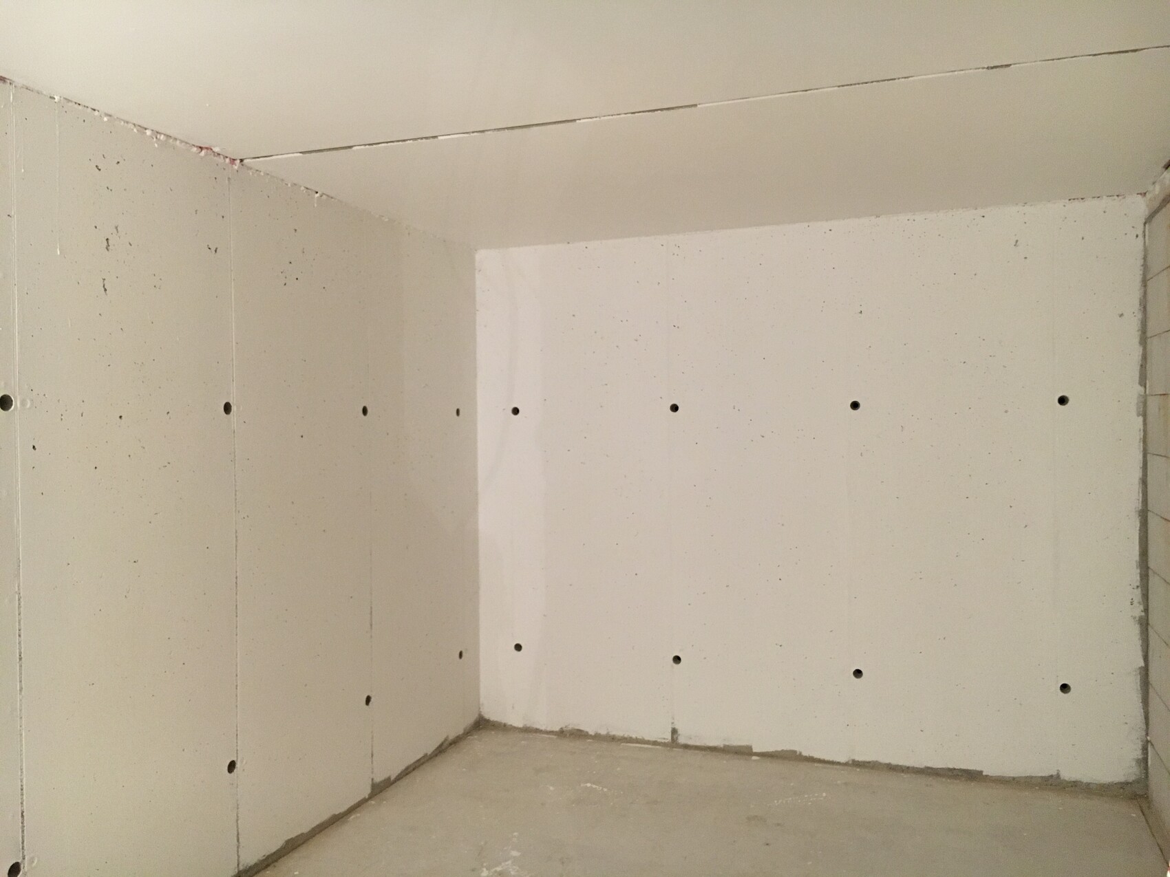 Leerer Raum mit unbehandelten Gipskartonplatten an Wänden, zahlreiche Löcher sichtbar.