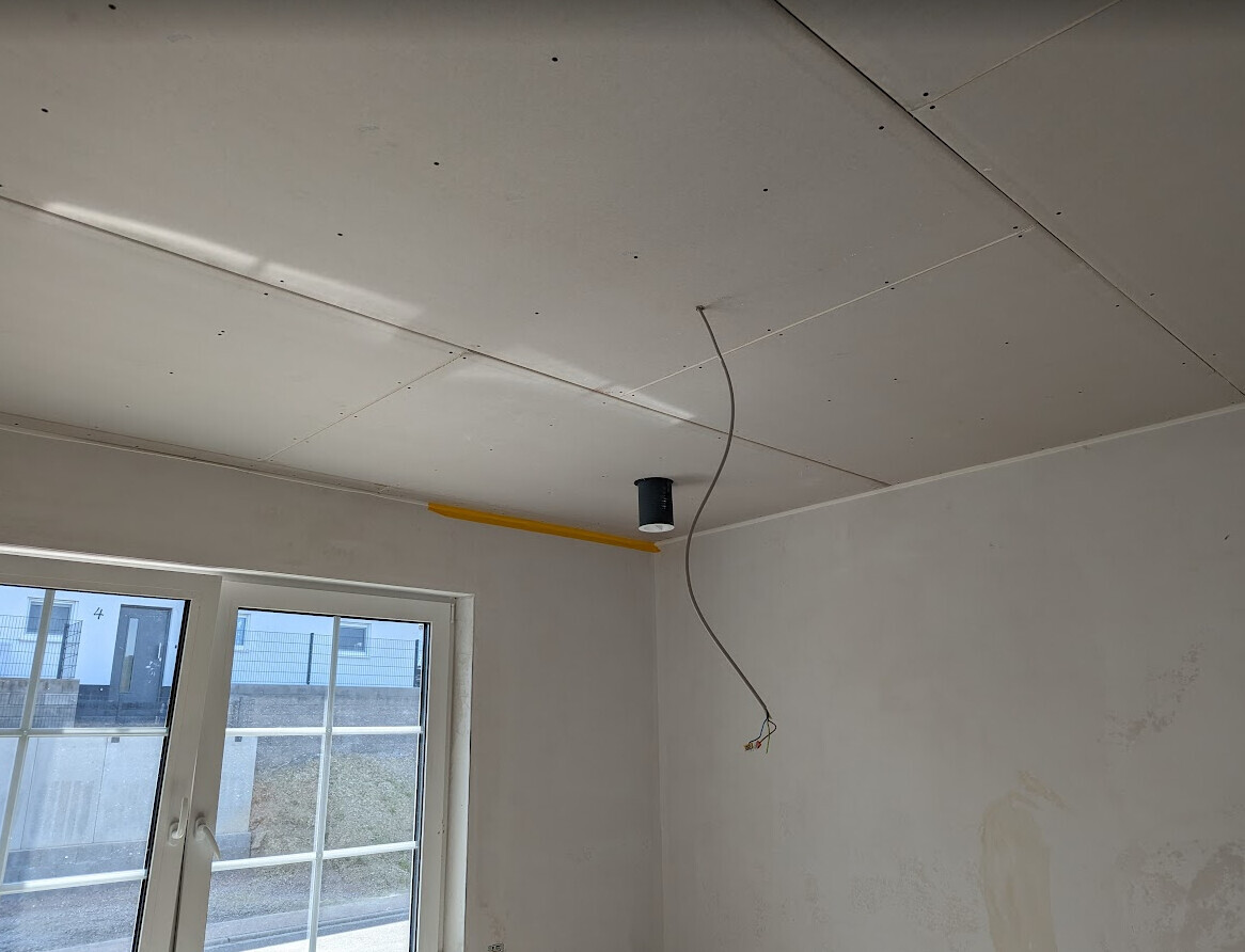 Unfertige Decke mit Gipskartonplatten, lose Kabel und eine hängende Leuchte neben dem Fenster.