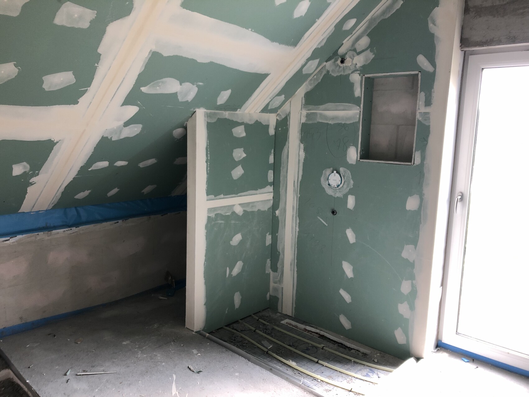 Unvollendeter Dachboden-Bau mit grünen Gipsplatten, schräger Decke, Tür aus Glas und Bauarbeiten.