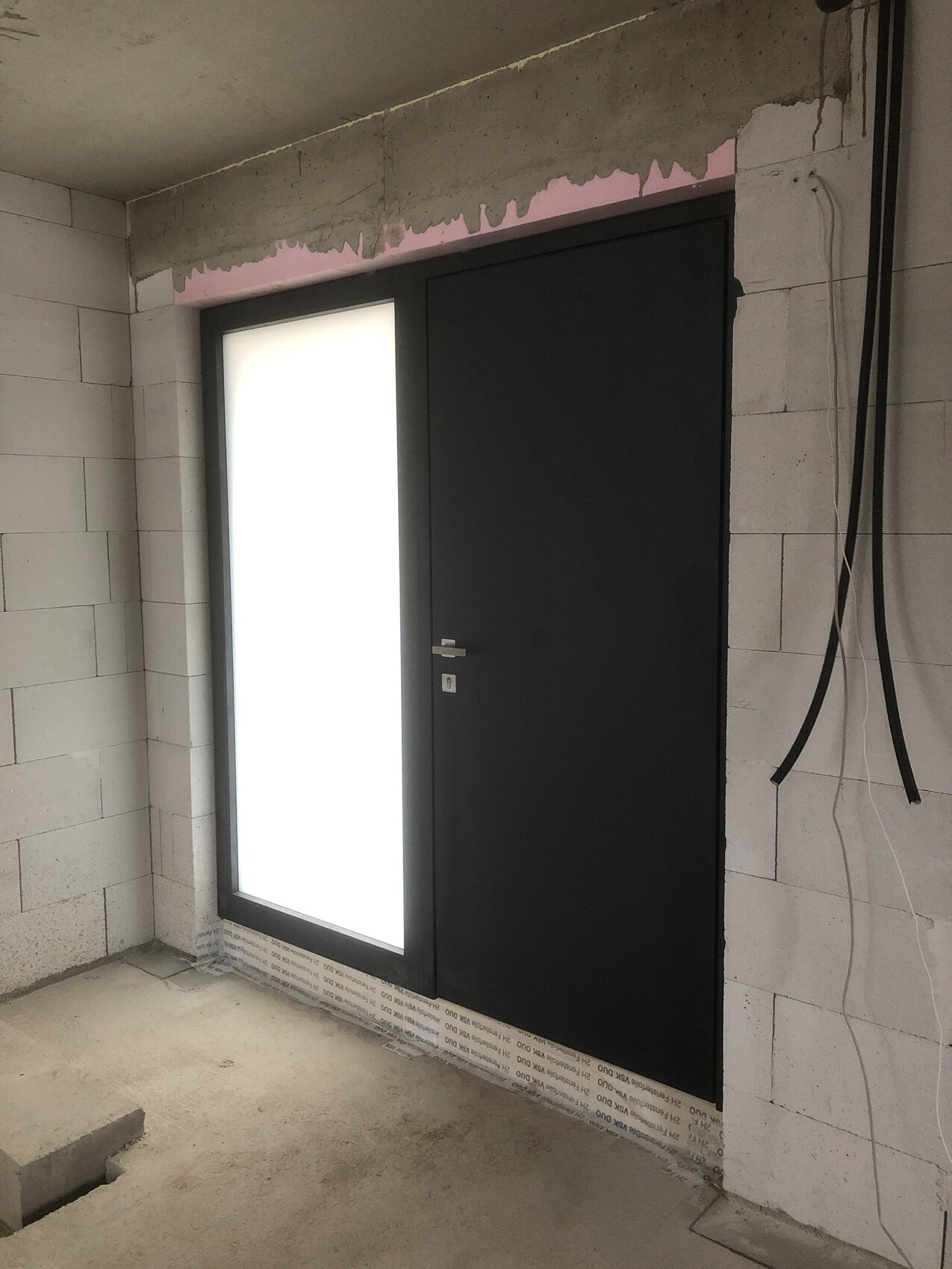 Rohbau: dunkle Tür neben milchweißem Glasfenster; Kabel an Wand, unfertige Betonwände.