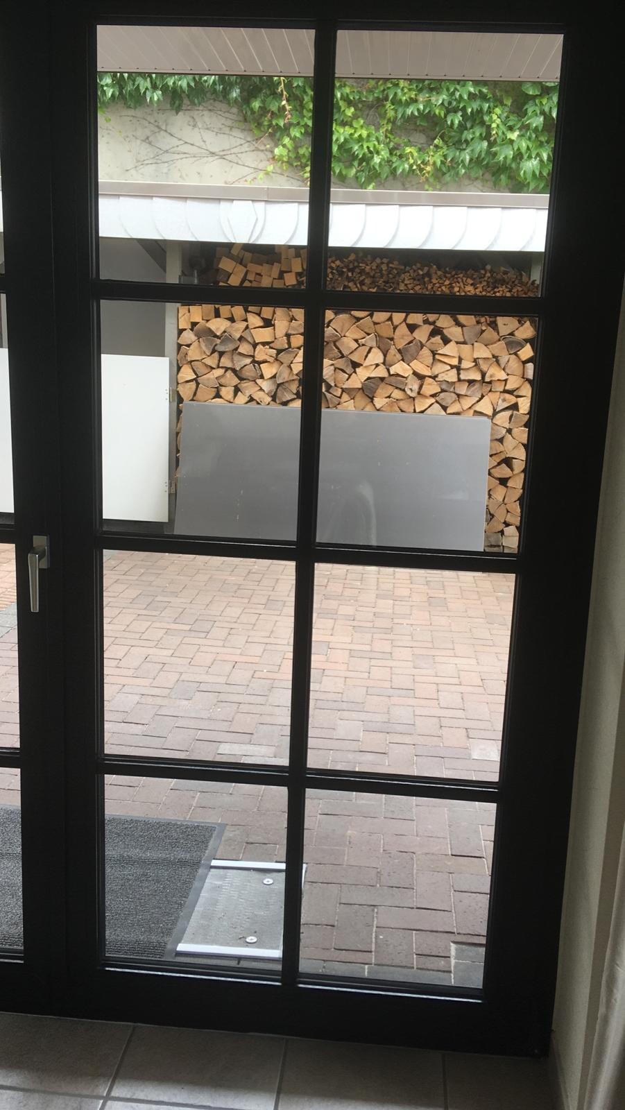 Gläserne Tür, Blick nach außen auf Holzstapel, Metallblech, gepflasterten Hof und Rankpflanzen