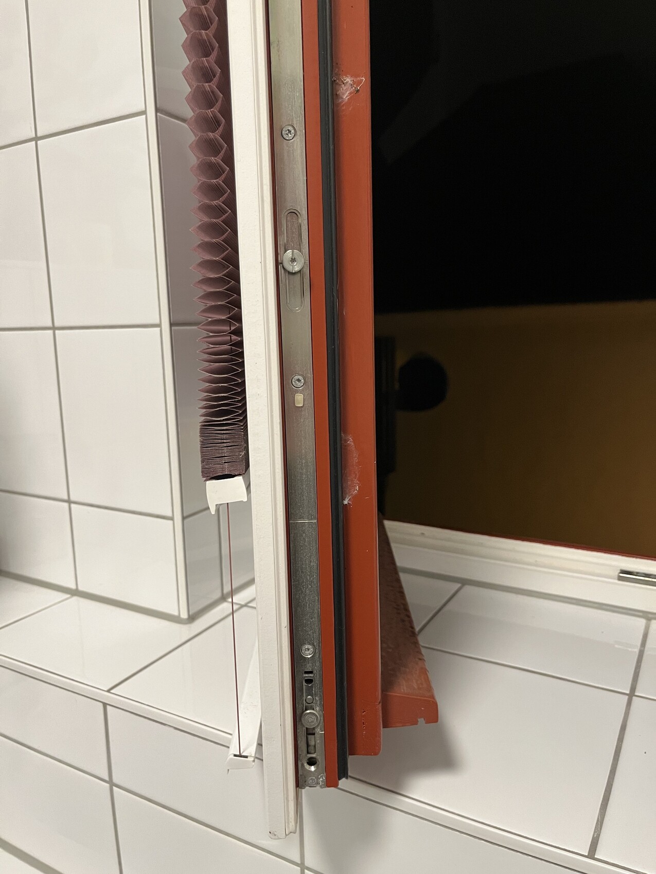 Offener Türrahmen aus rotem Metall mit Dichtung, brauner Falt-Jalousie und weißer Fliesenwand.