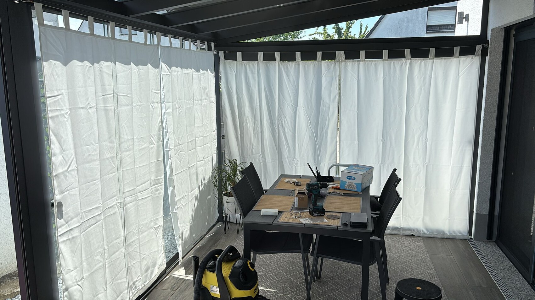 Überdachte Terrasse mit Esstisch, Stühlen, Werkzeugen und weißen Vorhängen