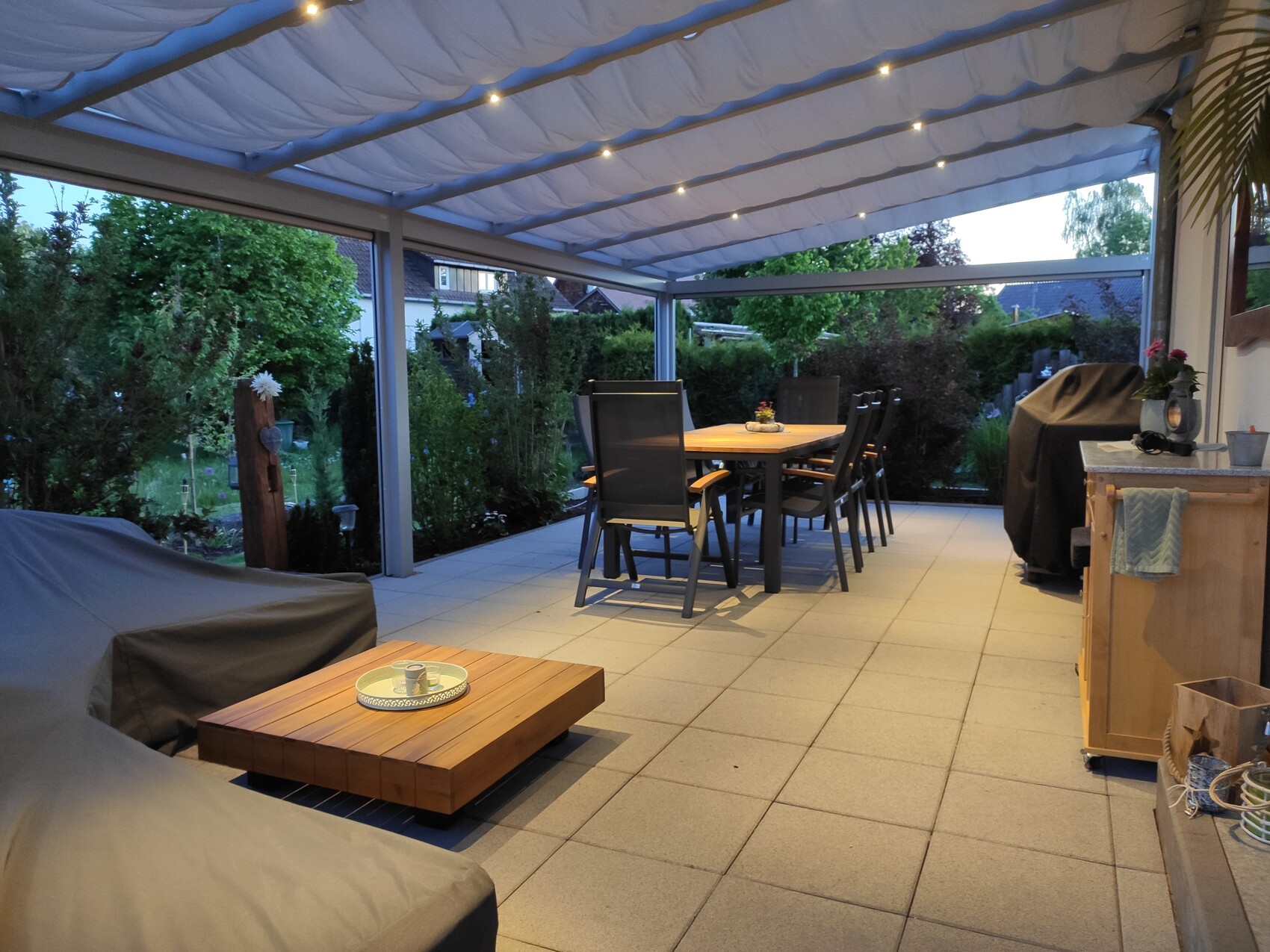 Überdachte Terrasse mit Esstisch und Stühlen, Garten im Abendlicht.