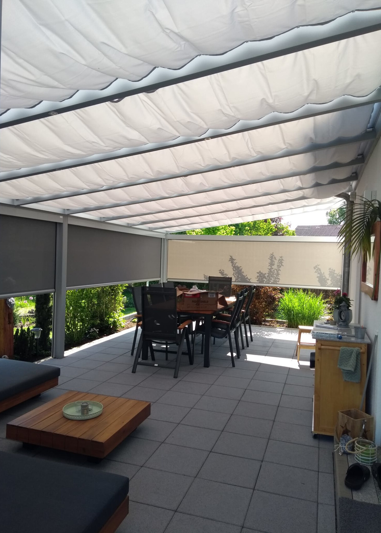 Überdachter Außenbereich mit rechteckigem Esstisch und Stühlen, grauen Fliesen und Gartenpflanzen.