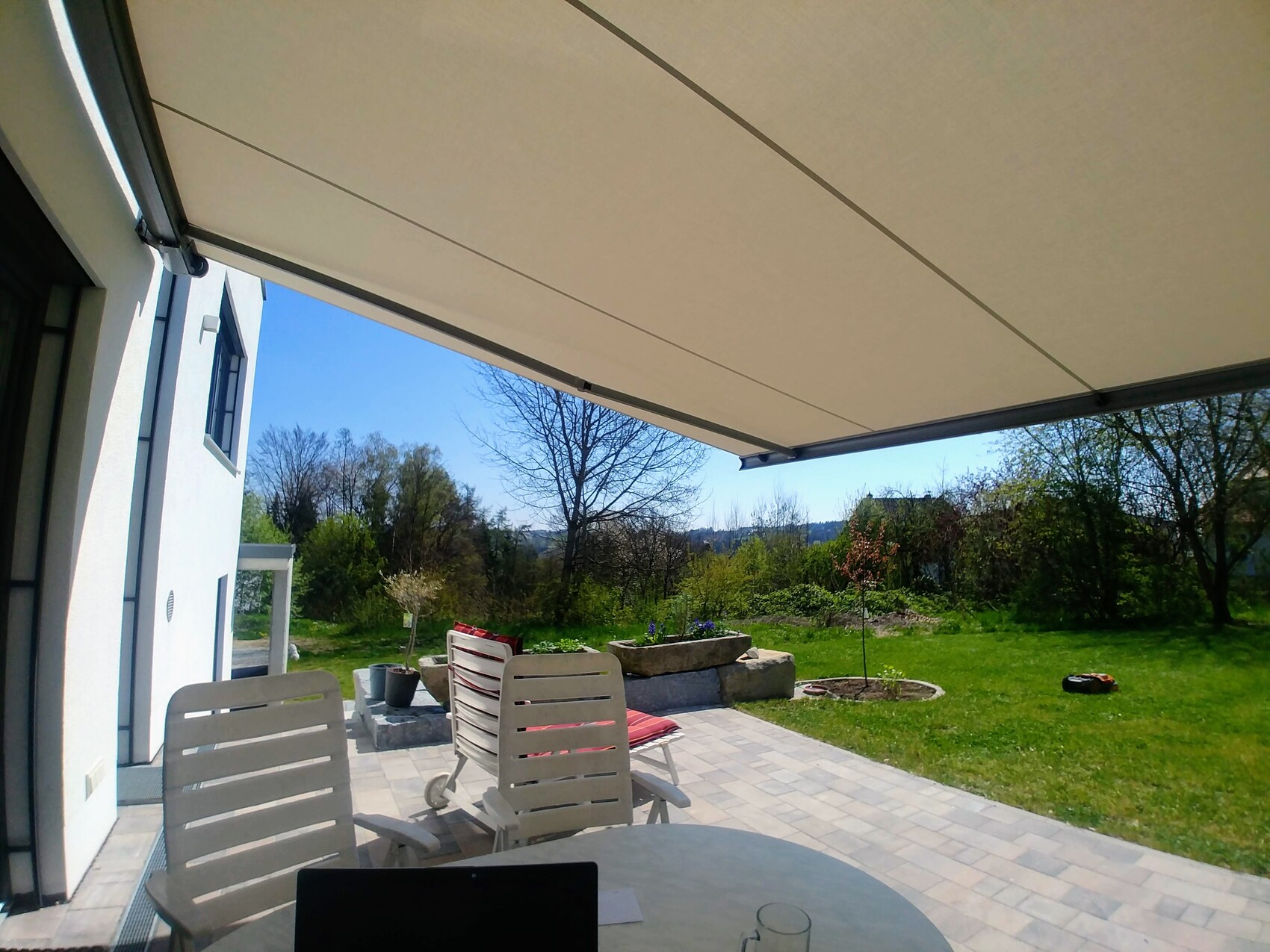 Terrasse mit ausziehbarer Markise, Gartenblick, weiße Fassade, Stühle und Tisch mit Laptop