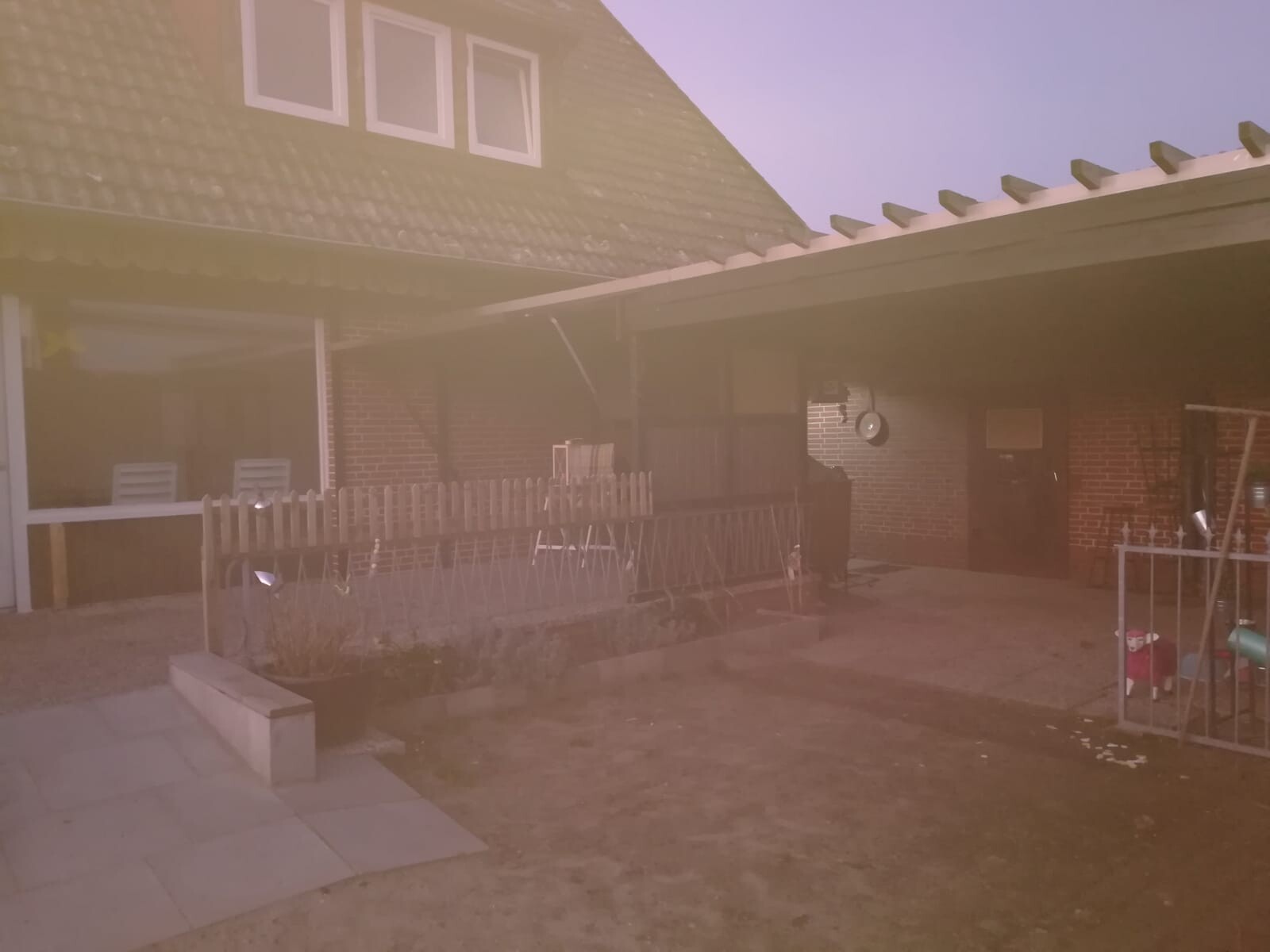Gartenbereich vor einem Haus mit überdachter Terrasse, Holzzaun und Spielzeug im Vordergrund.