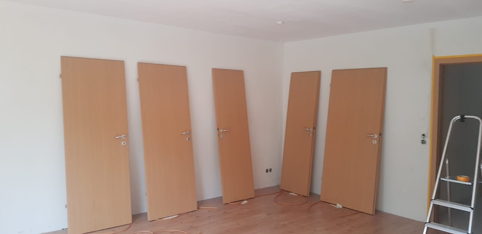 Raum mit fünf Holztüren, an der Wand gelehnt; Leiter und Kabel liegen auf dem Boden.