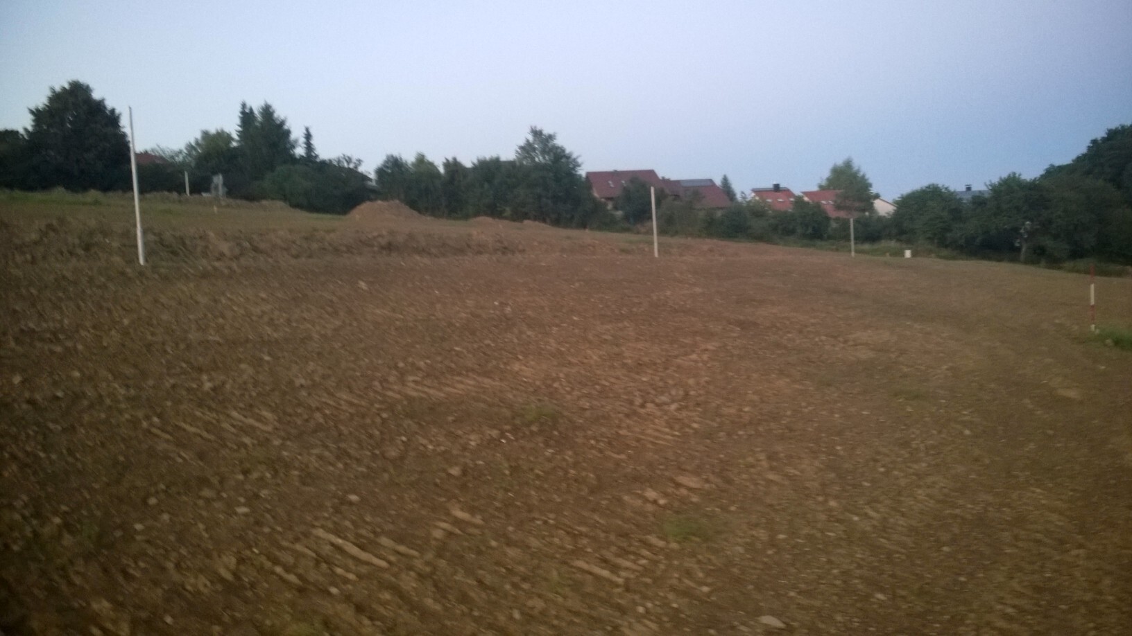 Unbearbeitetes Bauland mit Markierungsposten und Häusern im Hintergrund
