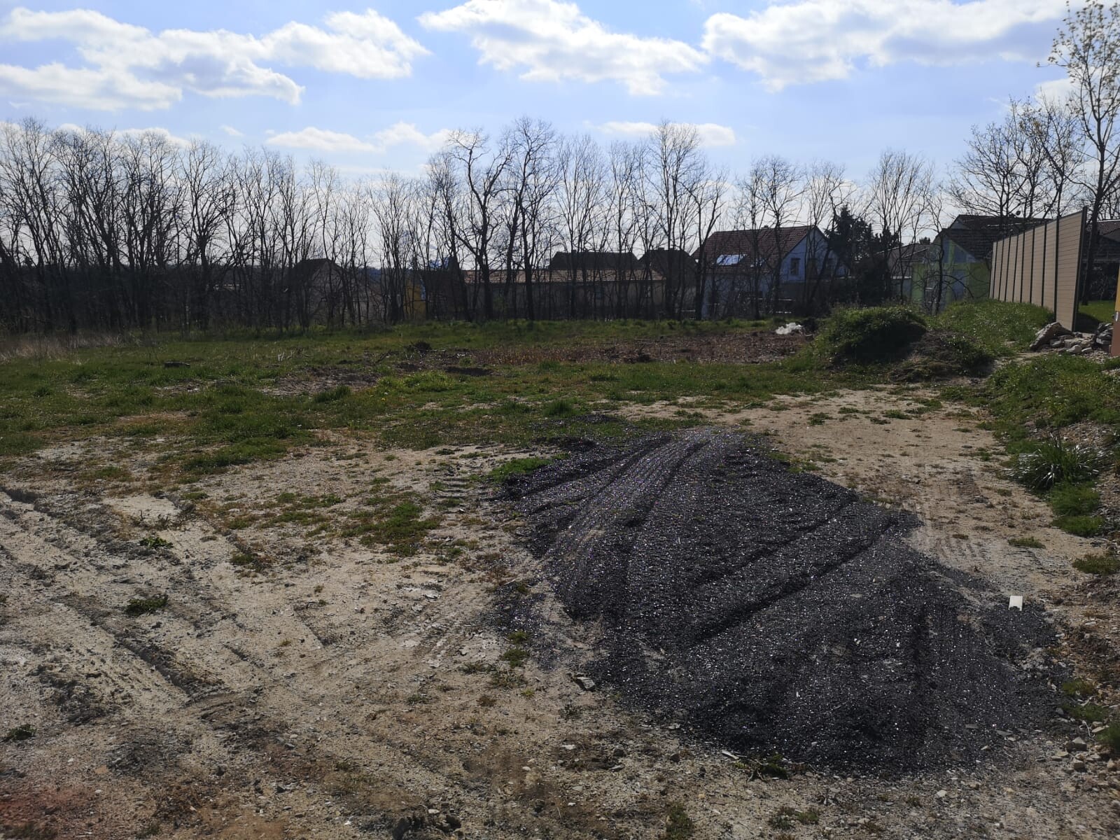 Leeres Baugrundstück mit dunklem Kieshaufen, Grasflächen, kahler Baumreihe und Häusern im Hintergrund.