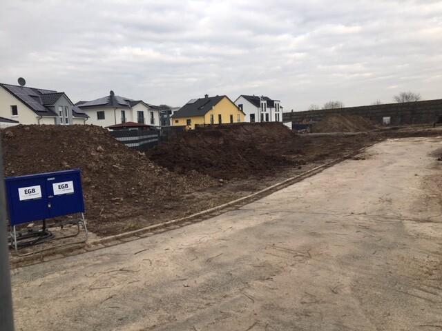 Baustelle in Wohngebiet: Große Erdhaufen, unbefestigte Straße, Häuser im Hintergrund.