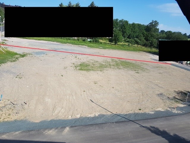 Leeres Baugrundstück mit roter Linie quer, Asphaltrand vorne, Bäume im Hintergrund.