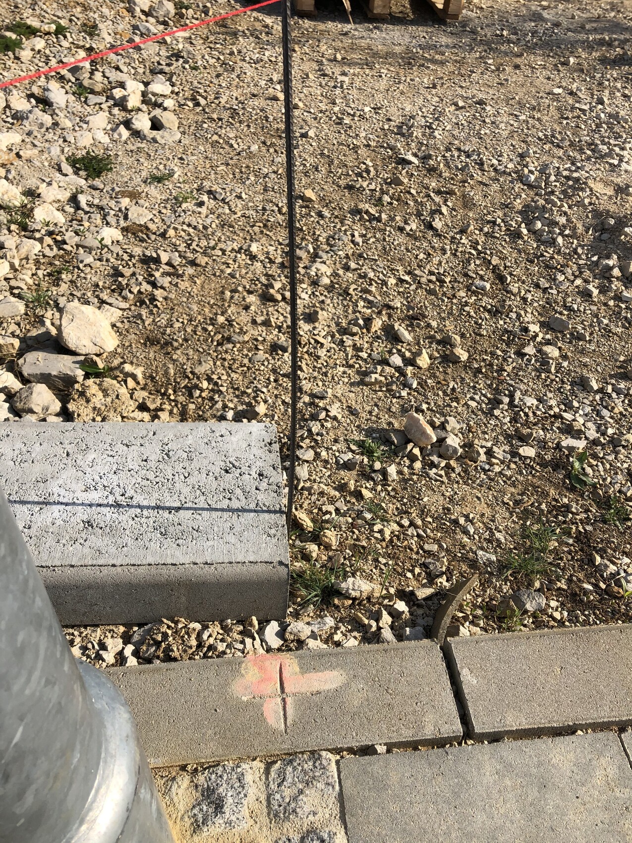 Baustelle mit Kiesboden, schwarzer Stab, pinkes Kreuz auf Bordkante, Betonblöcke daneben