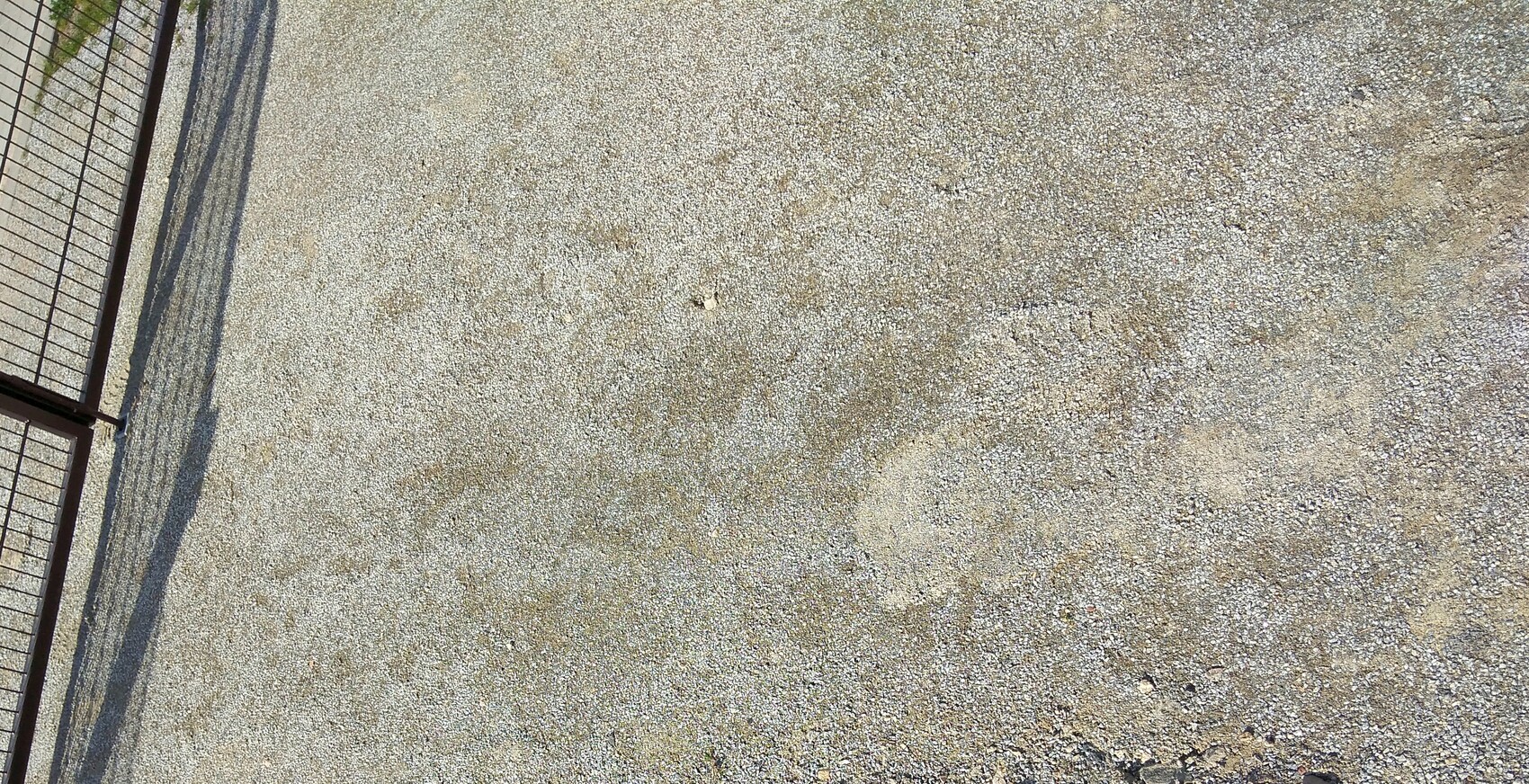 Schotterboden mit Metallzaun am linken Rand und langen Schatten des Zauns.