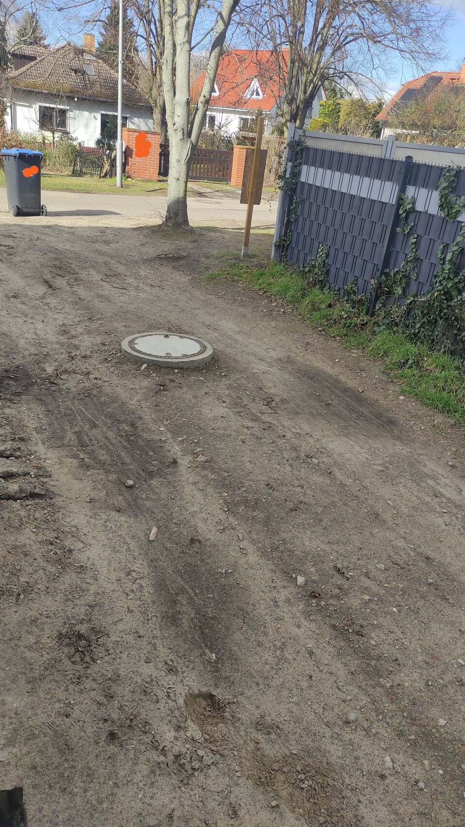 Unbefestigter Weg mit Mannloch, Mülltonne, Gartenzaun und Bäumen im Hintergrund.