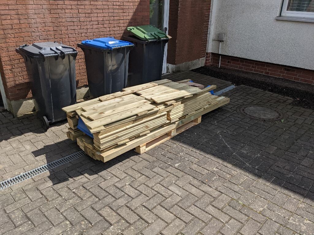 Holzlatten stapeln sich auf einer Palette neben Mülltonnen vor der Hausfassade.
