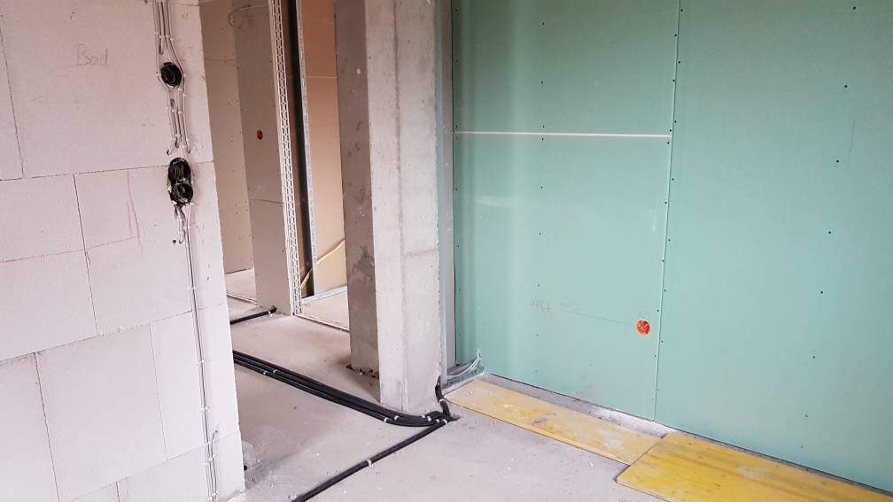 Baustelle: Innenraum mit grüner Trockenbauwand, Rohre, Betonstütze und offener Durchgang.