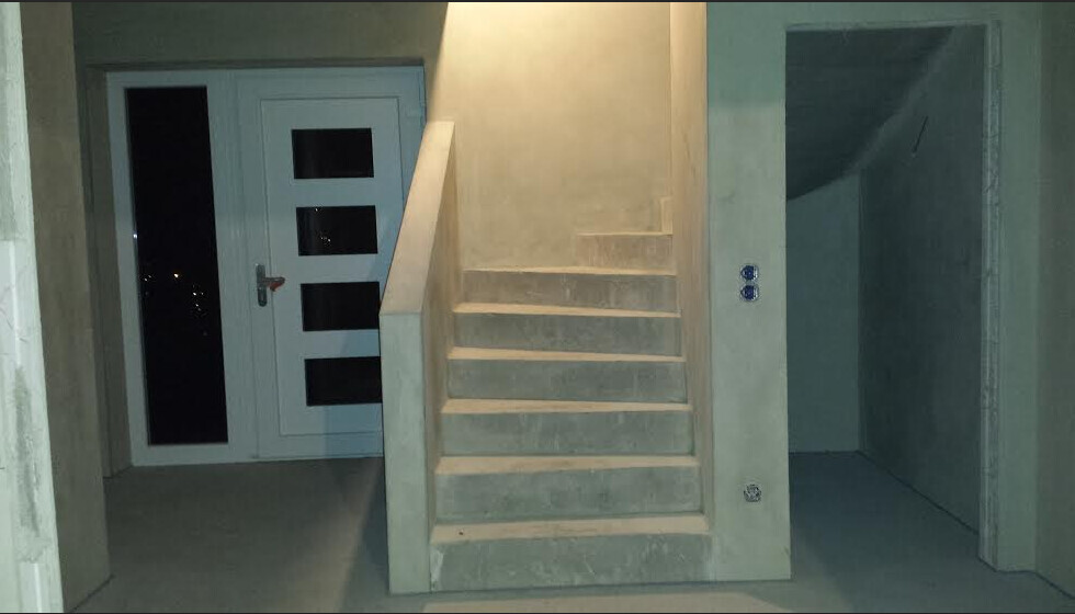 Unfertiger Innenraum: zentrierte Betontreppe, linke Tür mit Glasfenstern, rechter Durchgang.