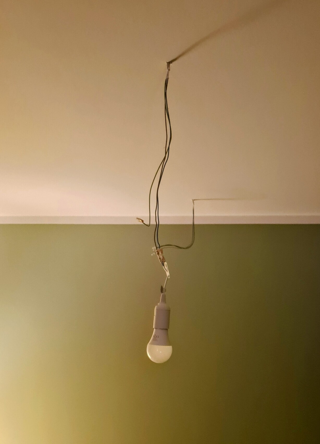 Hängende Glühbirne mit freiliegenden Kabeln von der Decke über einer grünen Wand.