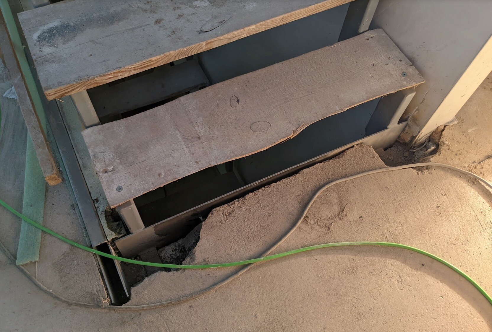 Holzstufen einer provisorischen Baustellentreppe, Staub, offener Spalt, grüne Leitung auf dem Boden.