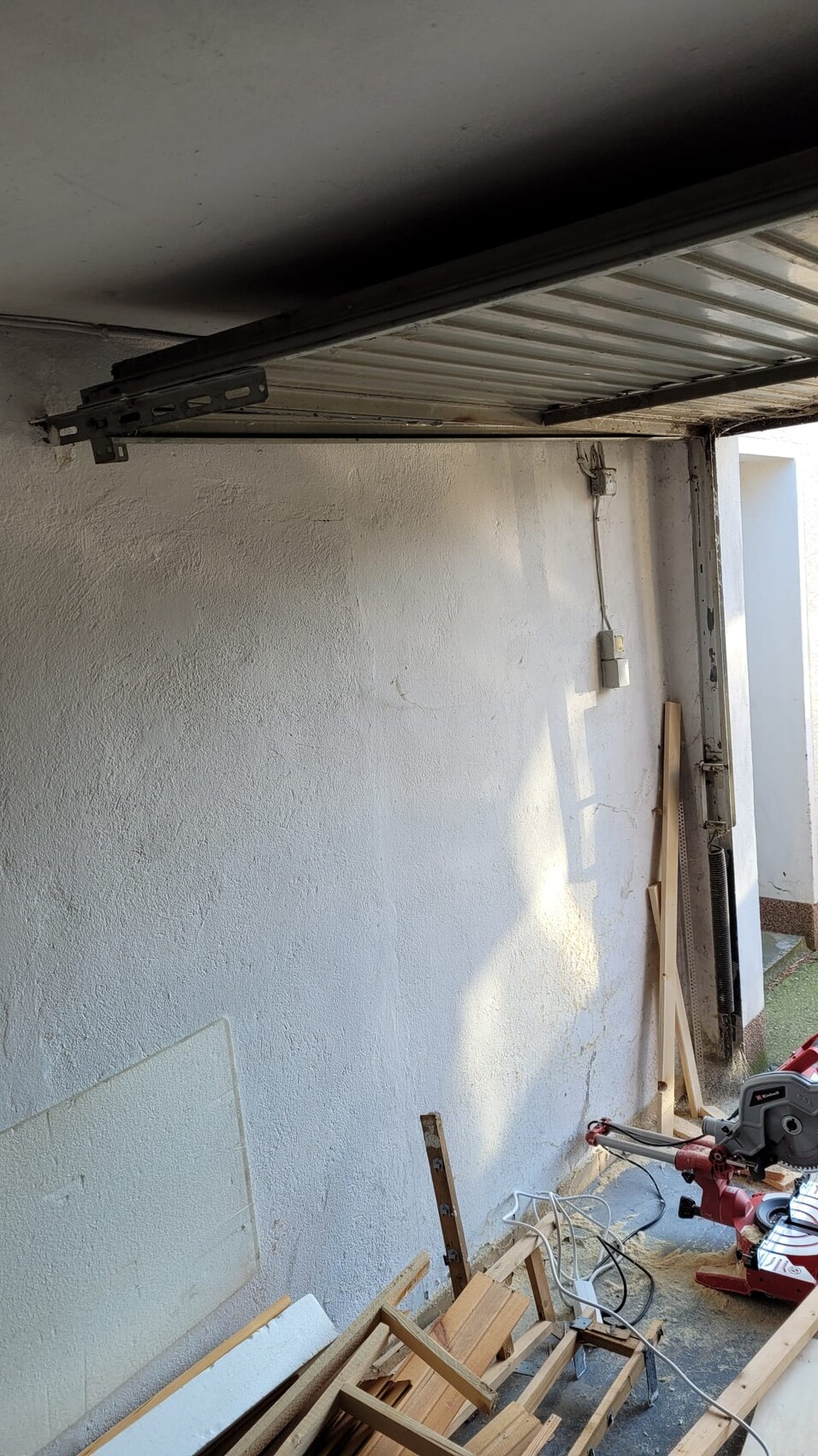Unordentliche Garage/Werkstatt mit halb geöffnetem Rollladen, Holzreste, Kabel und Kreissäge.