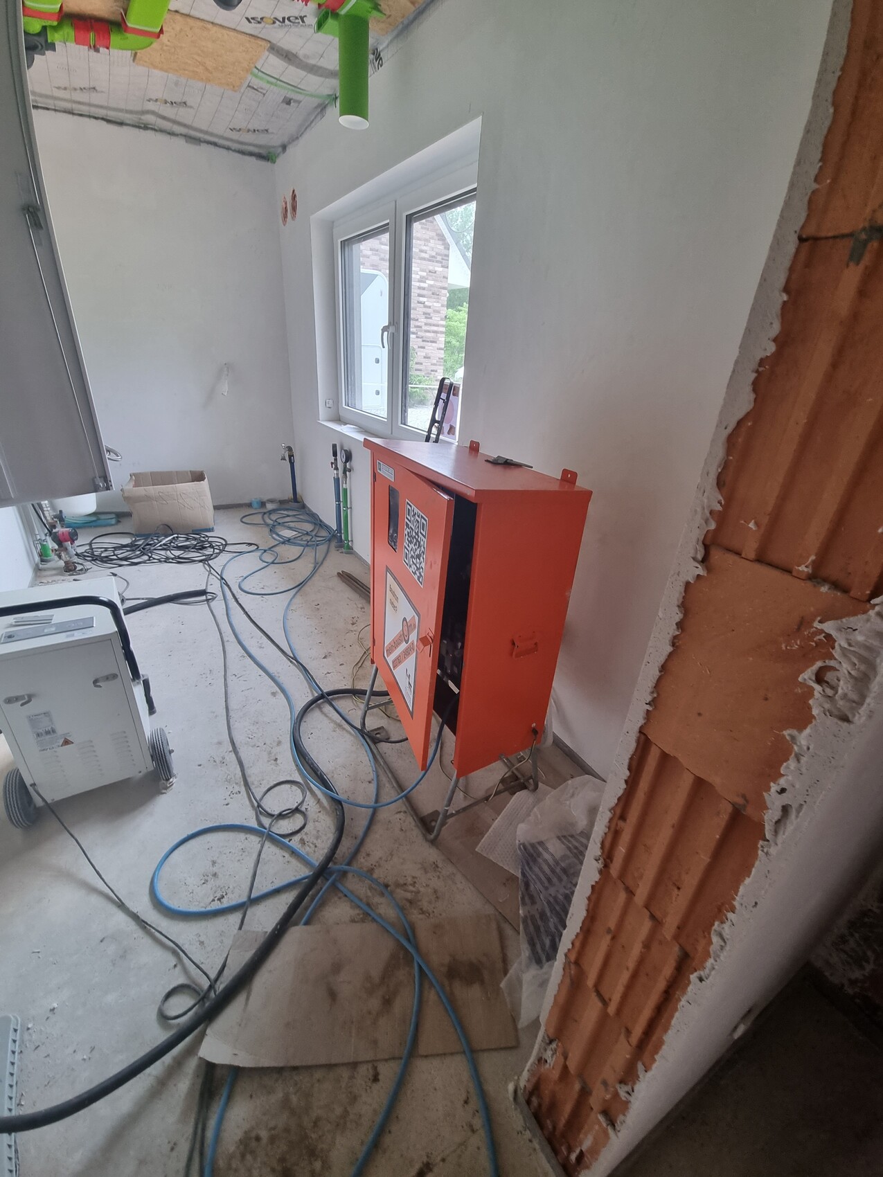 Baustelle: orangefarbener Schaltschrank, QR-Code, Kabelsalat, unfertige Wände, Fenster sichtbar.