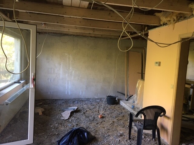 Unrenovierter Innenraum einer Baustelle: freiliegende Deckenbalken, lose Kabel, Schutt, Plastikstuhl.